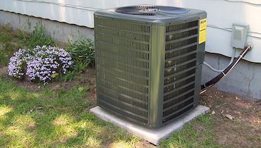 Central AC unit on concrete slab beside small flowering bush and patchy grass.