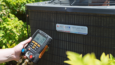 Close-up of hand holding digital HVAC manifold beside Aire Serv brand nameplate on outdoor central AC unit.