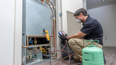 Male technician in disposable dust mask holding manifold gauge beside exposed HVAC unit.