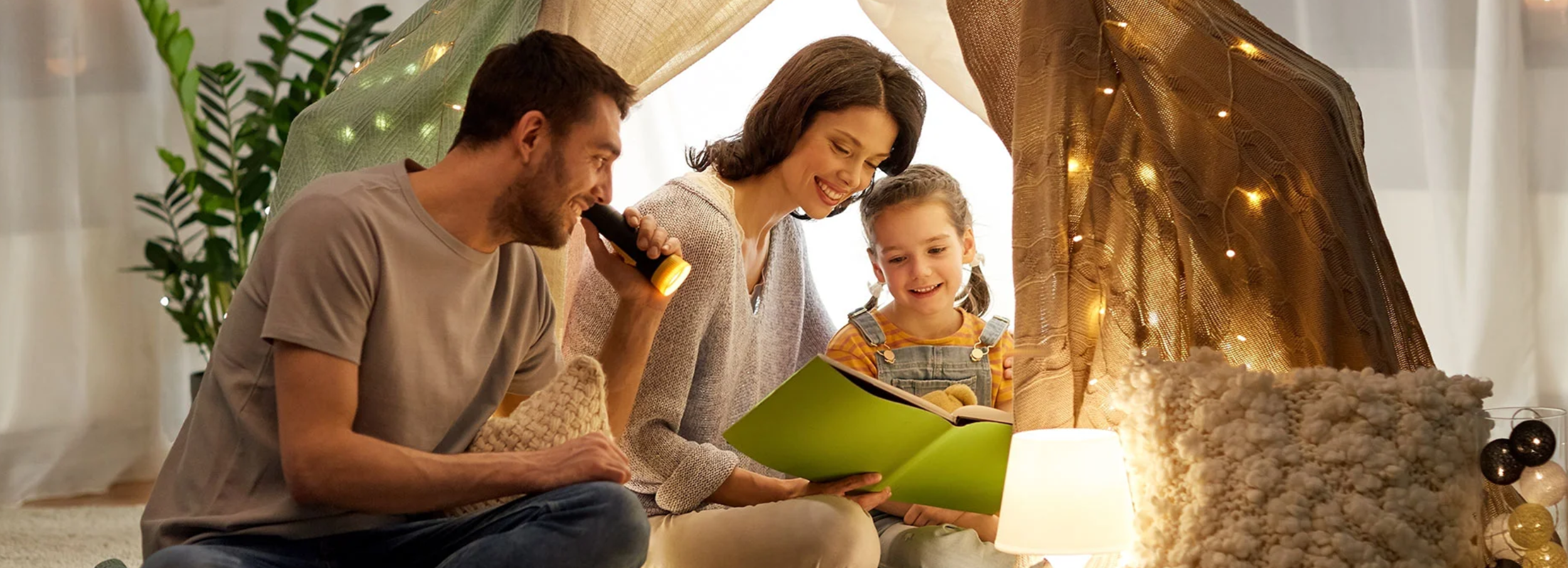 A man, woman, and young girl reading from a green book under a blanket fort.