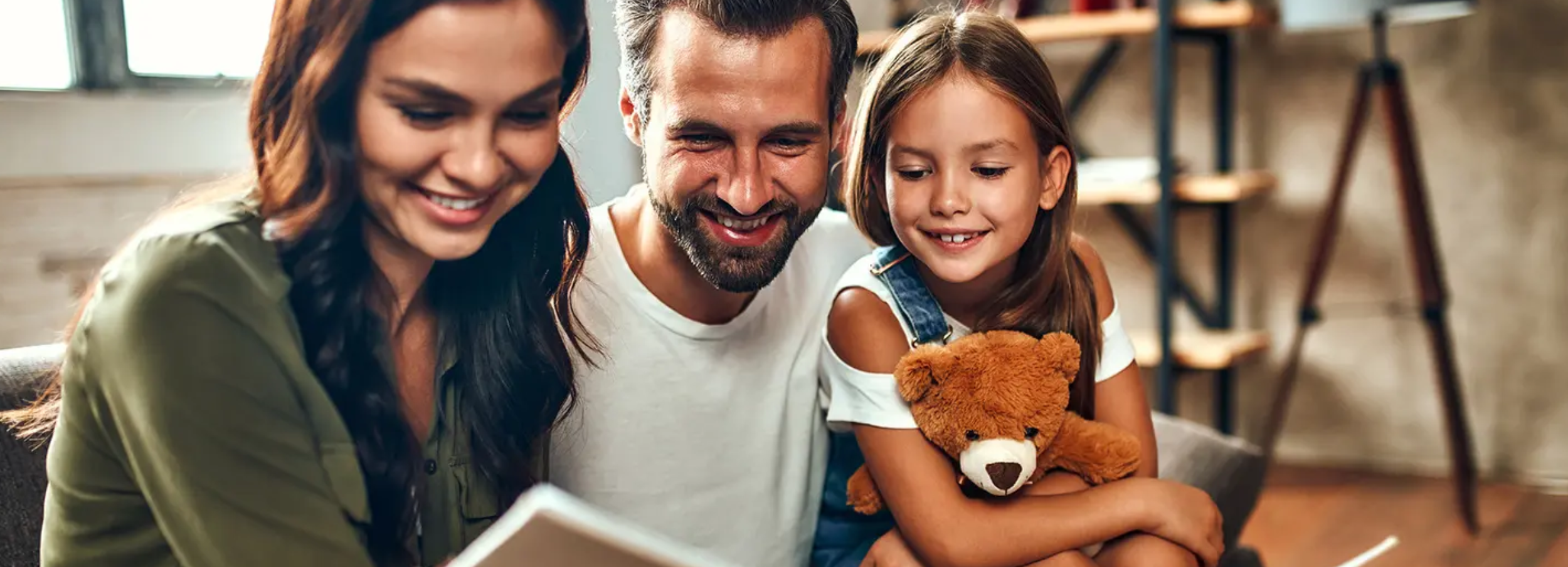 Parents smiling with young daughter holding a teddy bear.