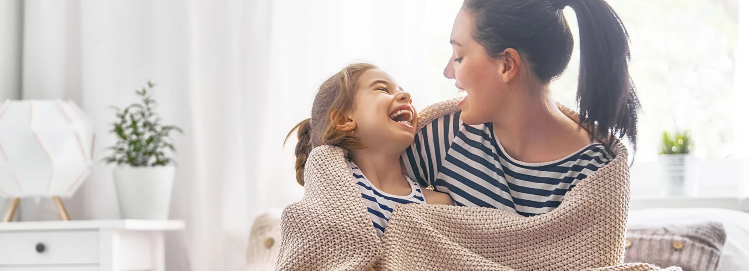 Smiling mother and daughter wrapped in woven blanket in brightly lit interior.